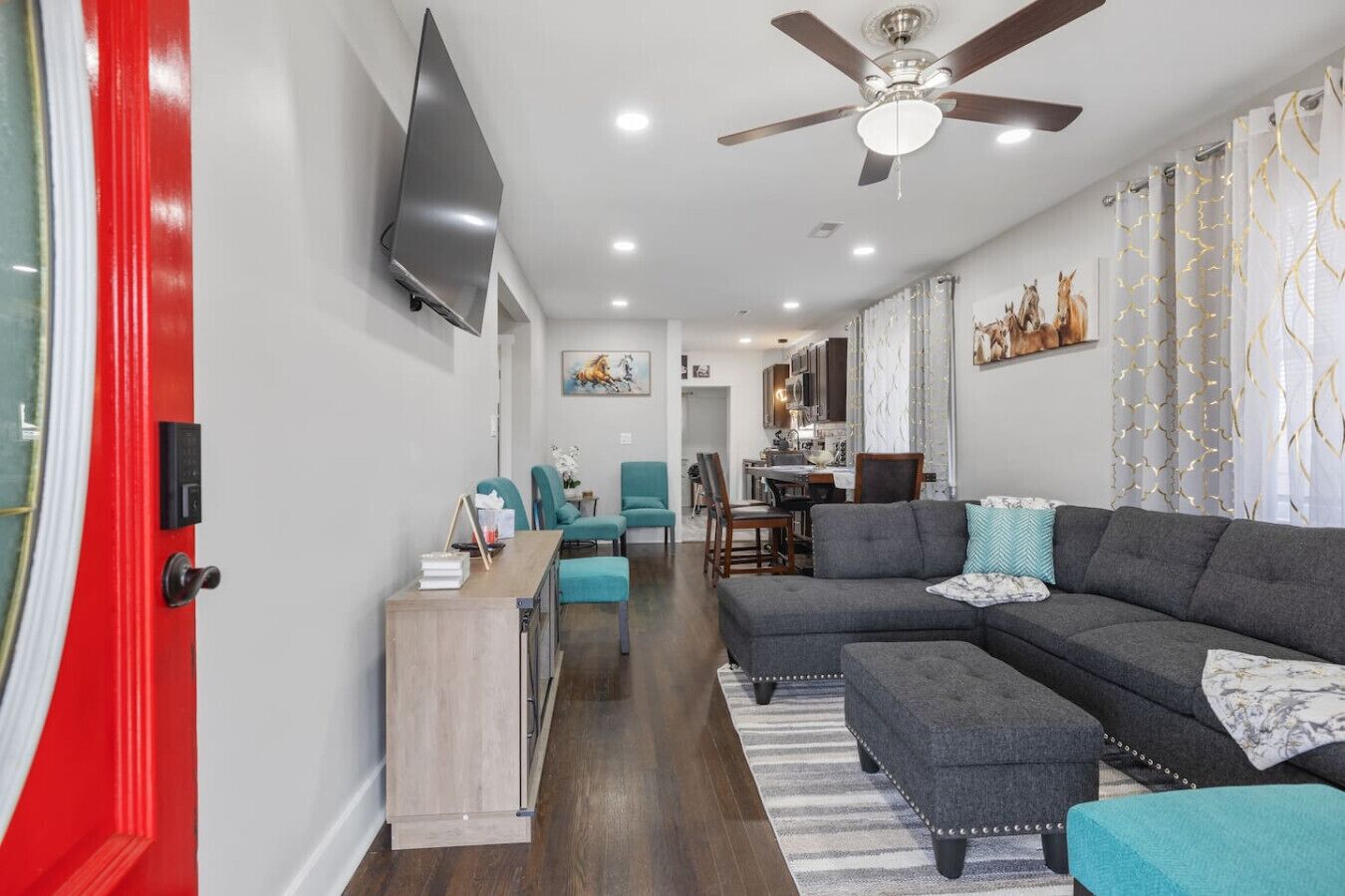 Un salon moderne avec un canapé d’angle gris, des fauteuils turquoise, une table basse en bois et un téléviseur à écran plat fixé au mur.