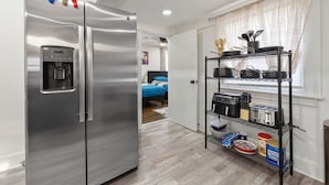 A compact kitchen with a stainless steel refrigerator, a coffee machine, and a black metal shelf unit holding kitchenware and appliances. - Spacious Bungalow Retreat Near Belle Isle, RVA! (Richmond)