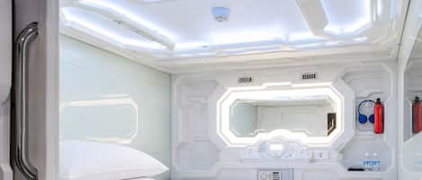 Una habitación futurista con una litera, un panel de control, un espejo y equipo de emergencia.