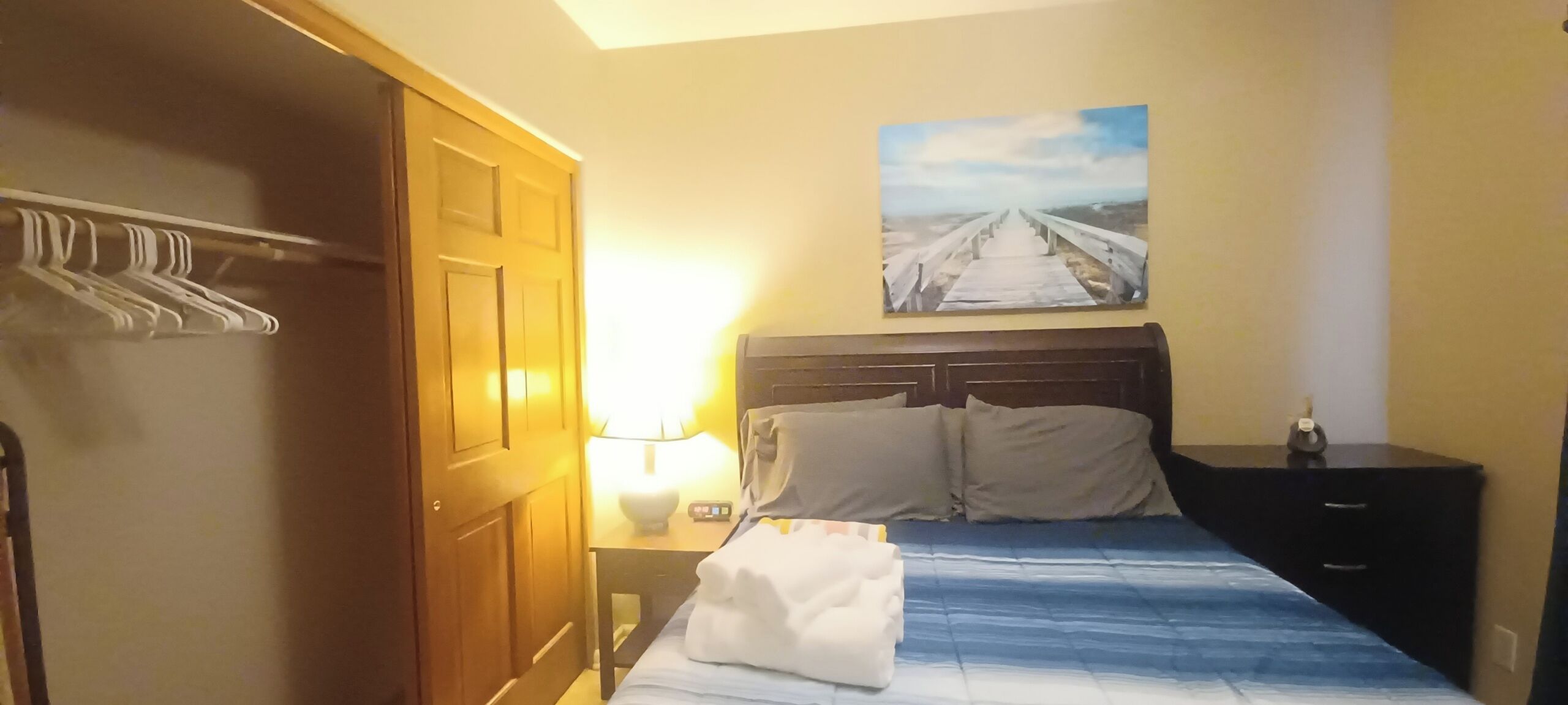 Hotellihuone, jossa on sänky, yöpöytä, lamppu, rantamaisemaa esittävä taulu ja puinen ovi.