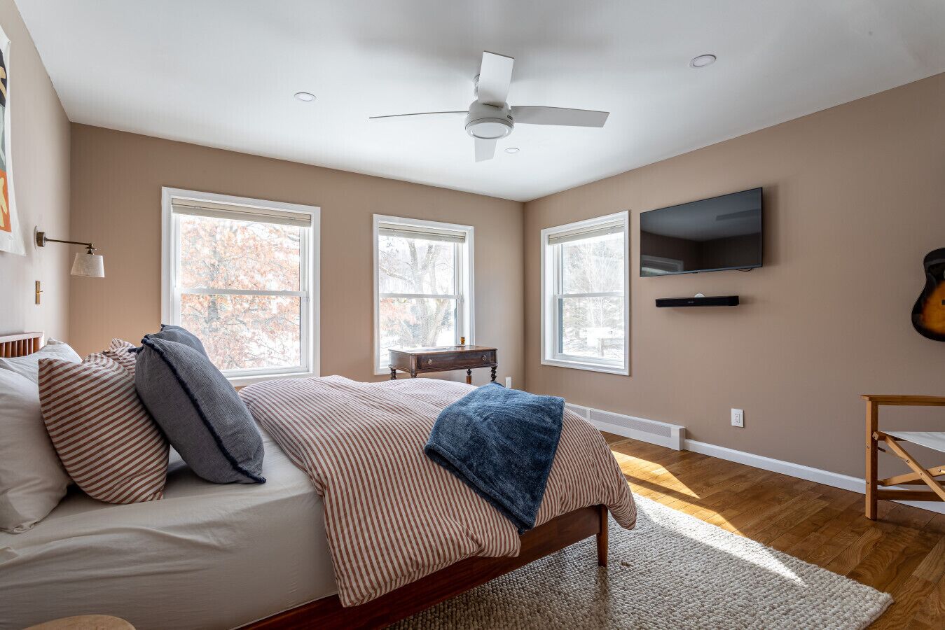 A bedroom with a bed, a ceiling fan, a TV, and large windows.