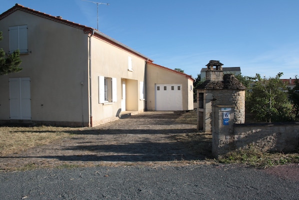 A building with a red-tiled roof, a stone structure with a blue sign, and a gravel driveway leading to a white garage door.