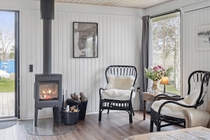 A cozy room with a wood-burning stove, two chairs, a table with a lamp, and a window with a view.