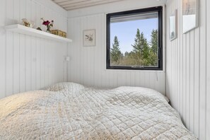 A bedroom with a bed, a window with a view of trees, a shelf with decorative items, and a framed picture on the wall.