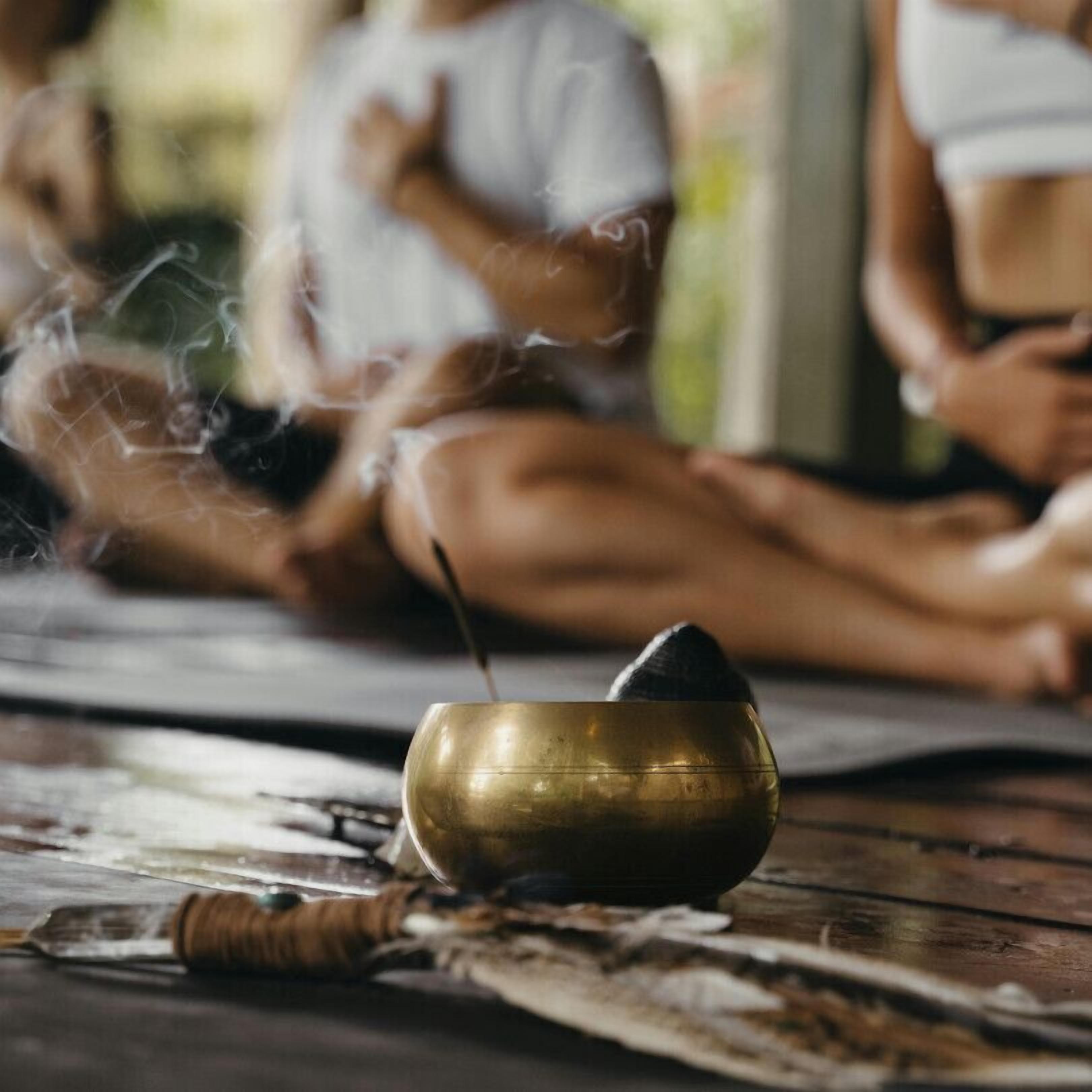 A meditation session with participants seated on mats, a brass bowl with a burning incense stick, and a wooden object on the floor.
