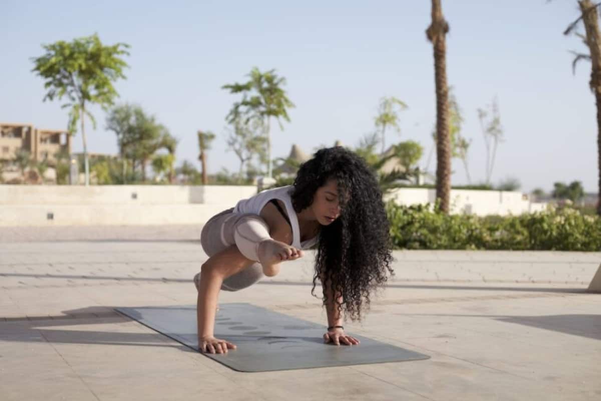 A person performing a yoga pose on a mat outdoors with palm trees and a building in the background.