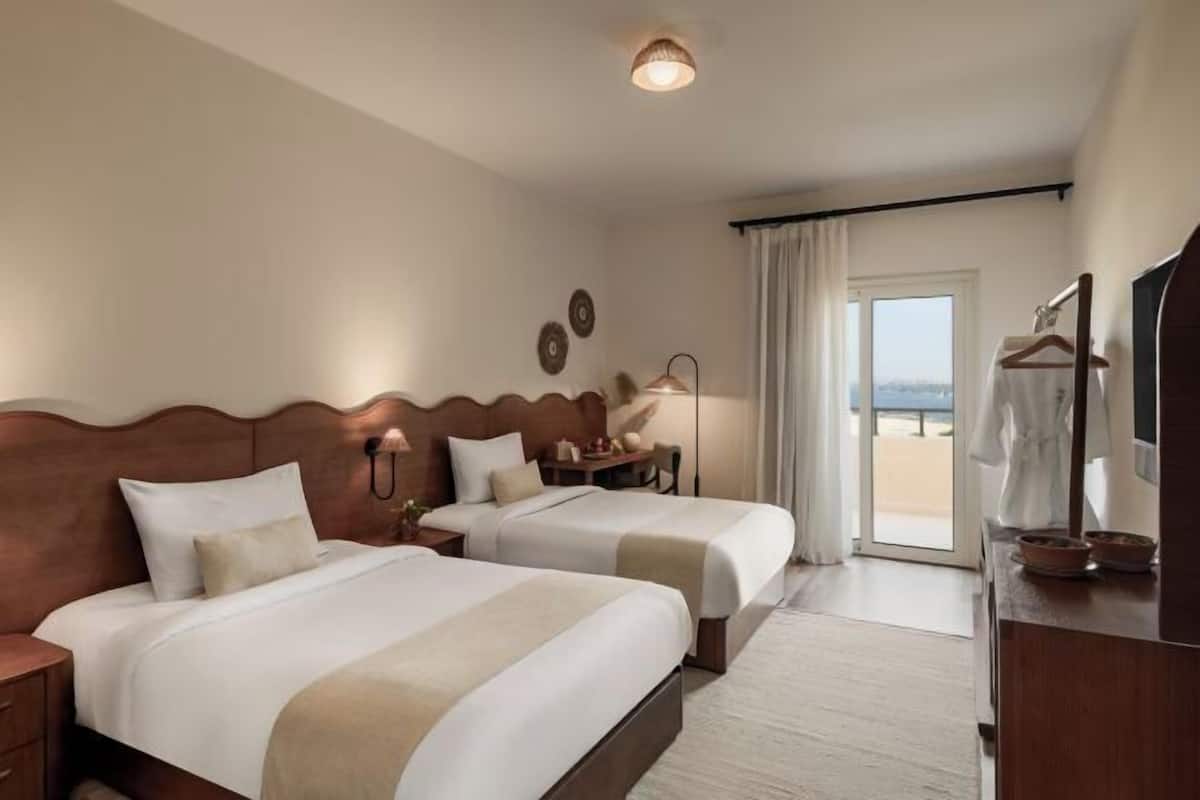 Habitación de hotel con dos camas, un escritorio, una silla, una lámpara, un armario y vistas al exterior.