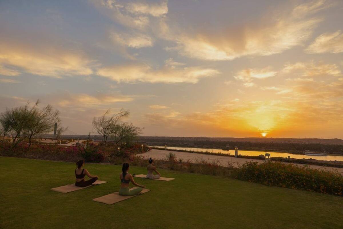 Three individuals meditating on yoga mats in a grassy area with a sunset view over a body of water.