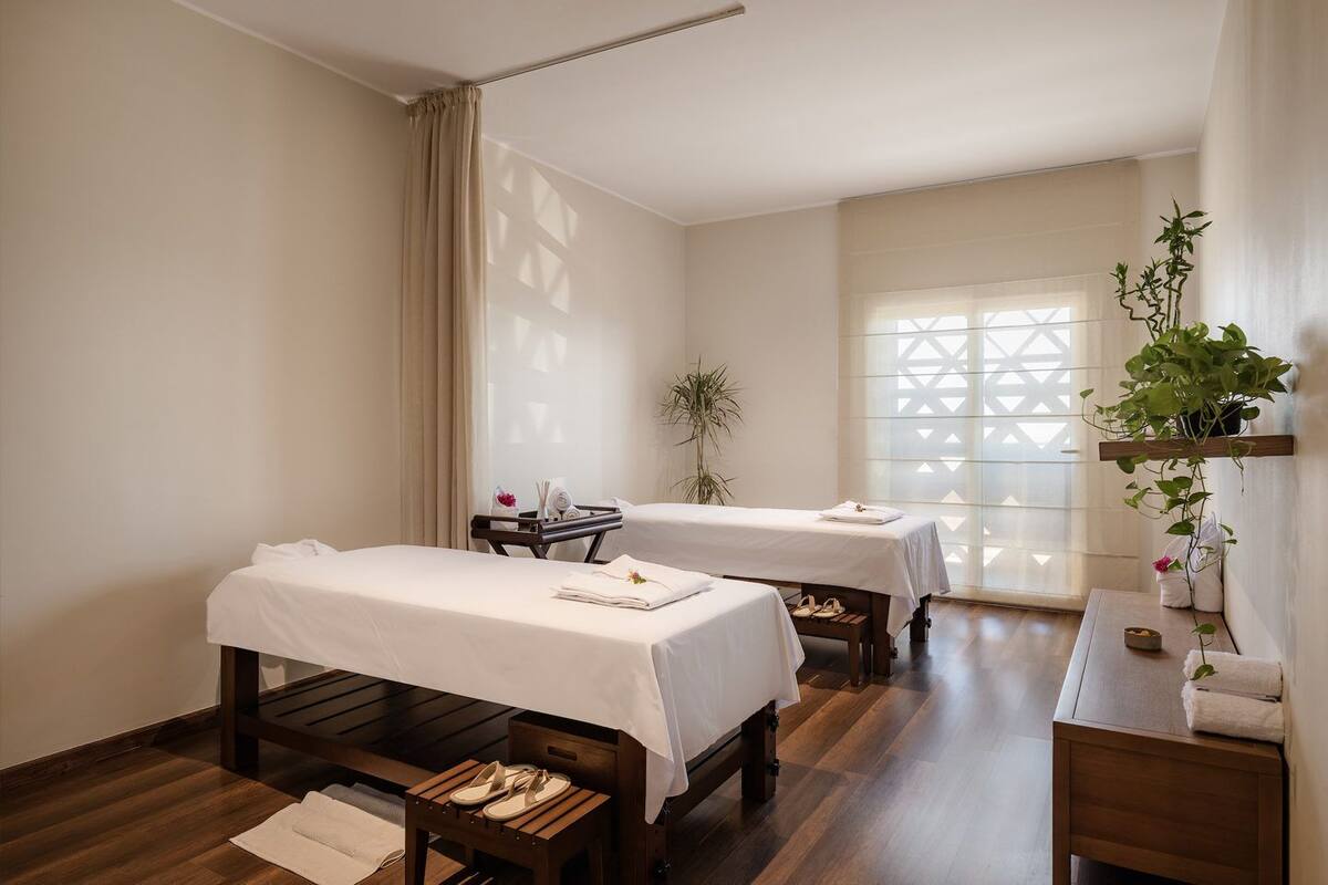 A room with two beds, a desk, a plant, and a window with a geometric pattern.