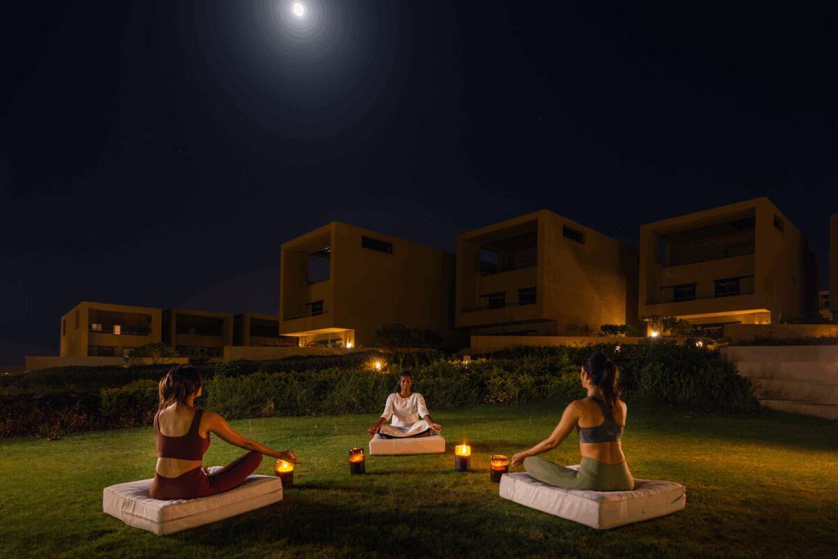 Three individuals meditating on outdoor cushions with candles, set against a backdrop of modern buildings and a night sky.