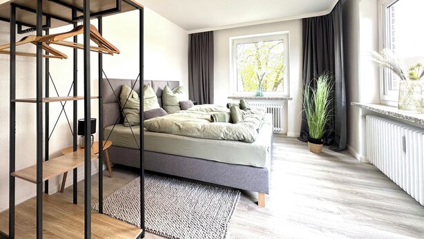 Quarto moderno com cama, guarda-roupa de madeira, radiador, janela com cortinas e uma planta.