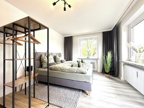 A modern bedroom with a bed, a wooden wardrobe, a radiator, a window with curtains, and a plant. - Modern apartment in the center with parking space - Hermannliving 12 (Schneverdingen)