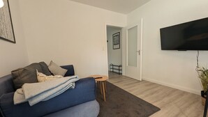 Uma sala de estar com um sofá azul, uma mesinha redonda, um tapete escuro, uma televisão de tela plana na parede e uma porta que dá para outro cômodo.