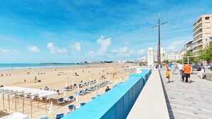 Une plage avec des chaises longues et des parasols, une jetée et, au loin, un beffroi.