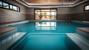 Une piscine intérieure moderne avec des parois en verre transparent, une terrasse en bois et une vue sur l’extérieur.