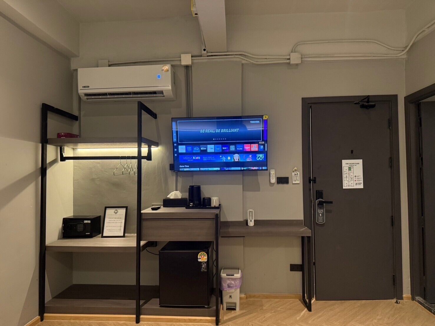 A hotel room with a TV displaying a streaming service, a desk with a coffee machine, a mini-fridge, and a door with a fire escape plan sign.
