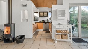 Ett modernt kök med en vedeldad spis, vita köksskåp och utsikt över trädgården genom en glasdörr.