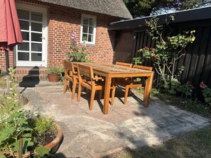 Uma casa de tijolos com telhado de palha, uma mesa e cadeiras de madeira e um jardim com vasos de plantas e flores.
