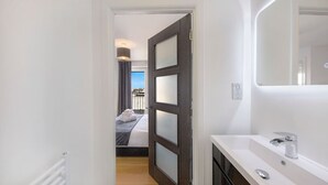 Un couloir étroit mène à une chambre à coucher avec un lit, une salle de bain avec un lavabo et un miroir, et une fenêtre donnant sur l’extérieur.