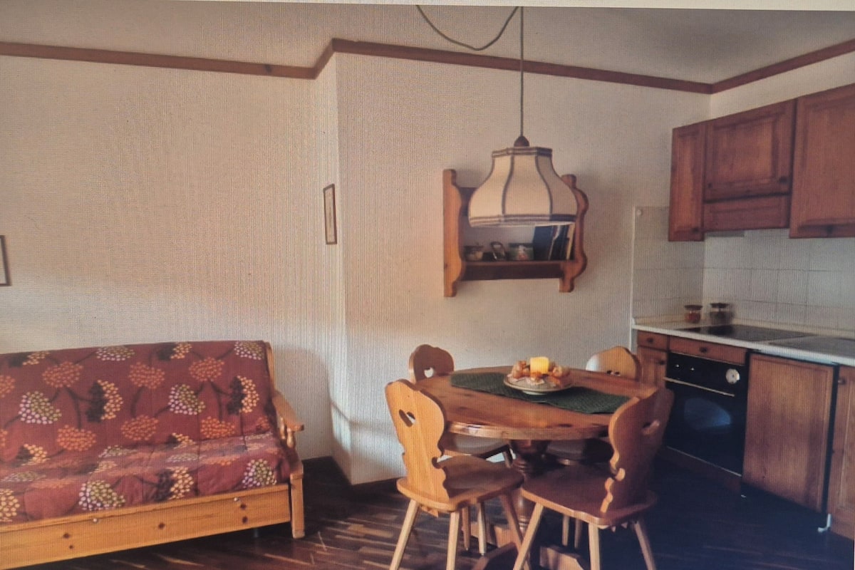 A compact kitchen with wooden cabinets, a dining table with chairs, a patterned sofa, and a hanging light fixture.