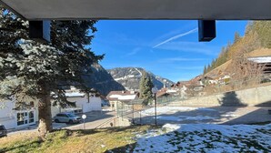 Vue depuis l’intérieur d’un bâtiment, donnant sur un paysage enneigé avec un arbre, un ciel dégagé et des montagnes au loin.