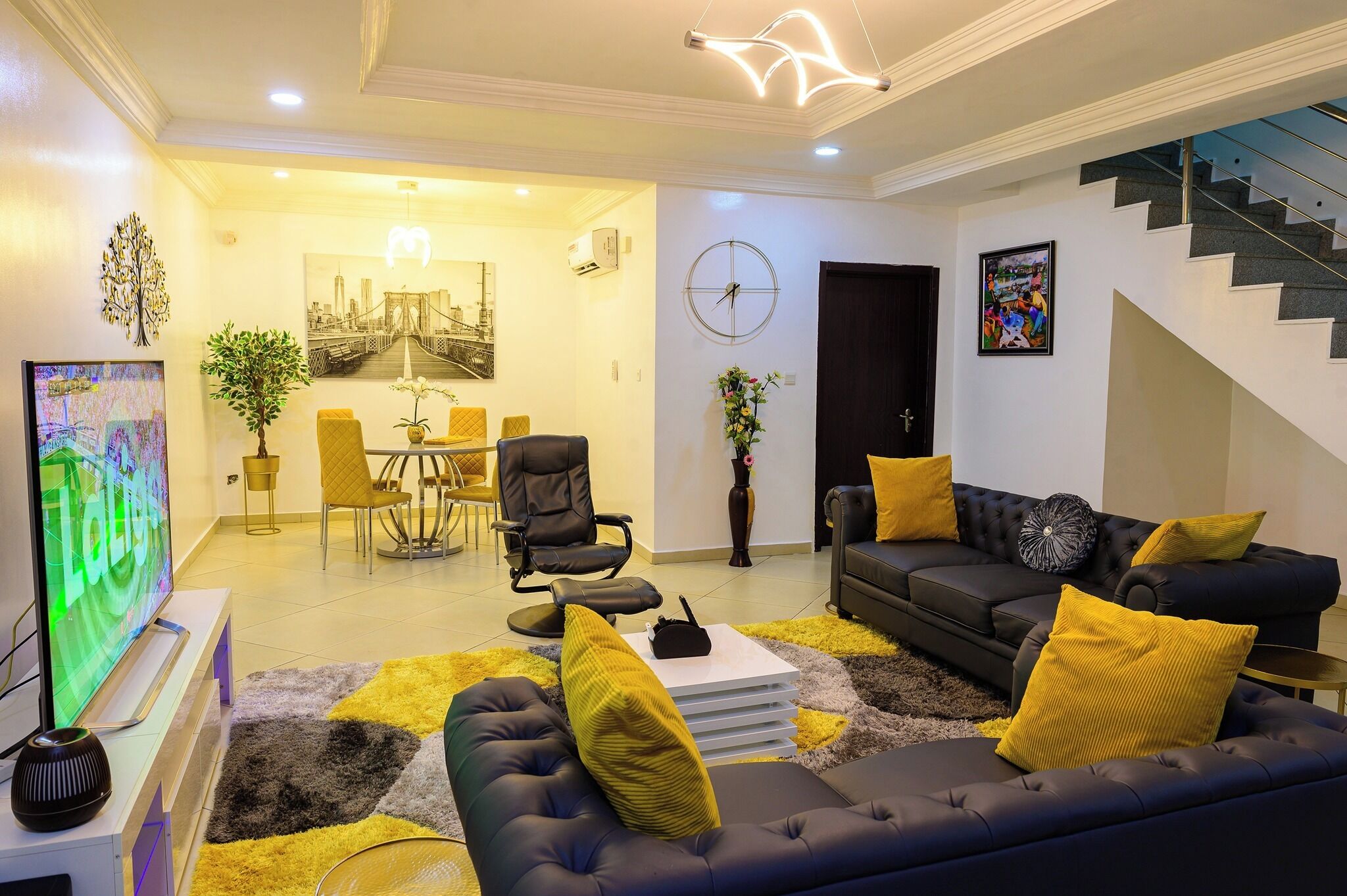 A modern living room with a black sofa, yellow cushions, a flat-screen TV, and a dining area with a table and chairs.