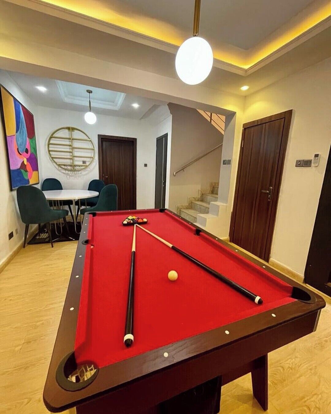 Una mesa de billar en una habitación con una escalera, un comedor y un cuadro abstracto y colorido en la pared.