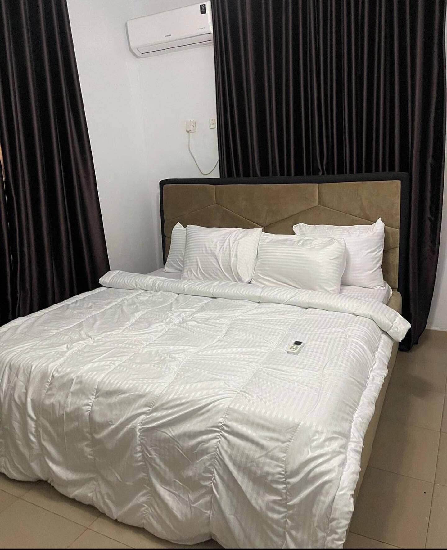 Una cama bien hecha con edredón y almohadas blancos, cabecero tapizado en color beige y aire acondicionado de pared.