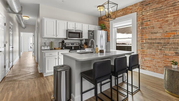 A modern kitchen with white cabinets, stainless steel appliances, a large island with bar stools, and a brick wall.