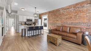 A modern kitchen with white cabinets, a central island with bar stools, a brown leather sofa, and a brick accent wall.