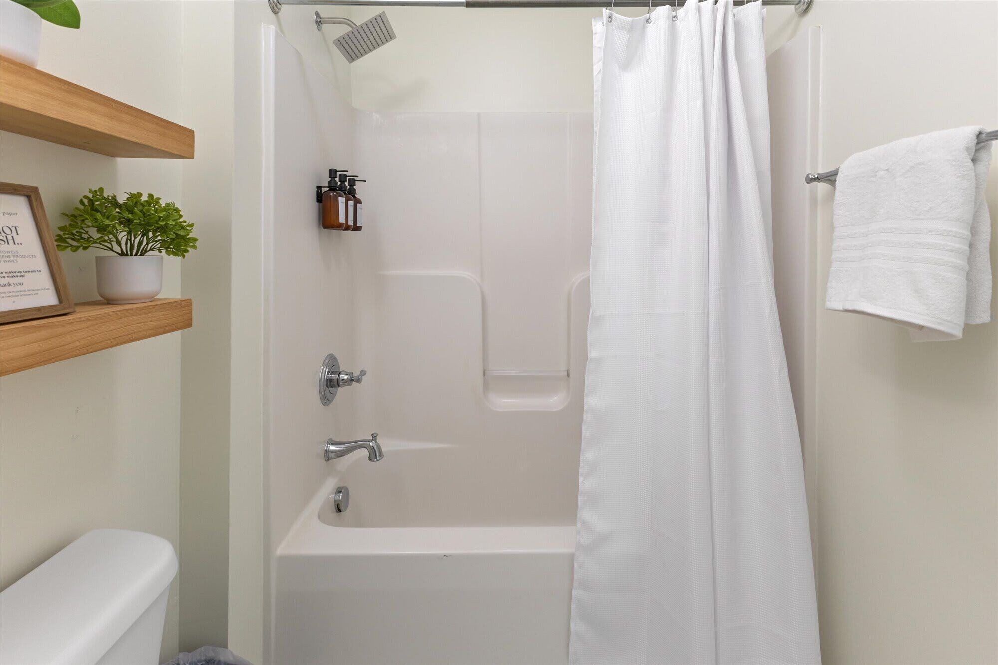 A bathroom with a bathtub, shower, white towel, and a shelf with a plant and a framed sign.