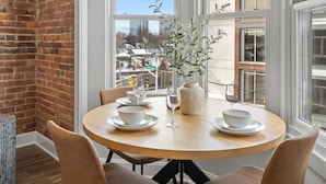 A dining area with a round table, four chairs, a vase with greenery, and a window with a view of buildings.