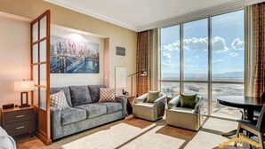 A modern living room with a large window offering a cityscape view, a gray sofa, a coffee table, and a small side table with a lamp. - MGM Signature-31-816 Jacuzzi Studio (Las Vegas)