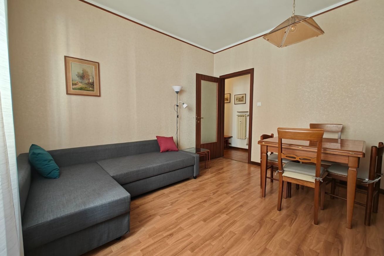 Ein Wohnzimmer mit einem grauen Ecksofa, einem Holzesstisch mit Stühlen, einer Stehlampe und einer Tür zu einem anderen Raum.