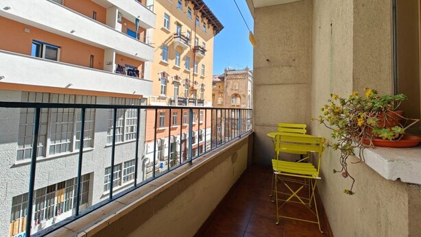 A balcony with yellow chairs and a potted plant, overlooking a cityscape with multi-story buildings.