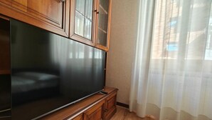 A room with a large flat-screen TV on a wooden cabinet, a window with sheer curtains, and a wooden floor.