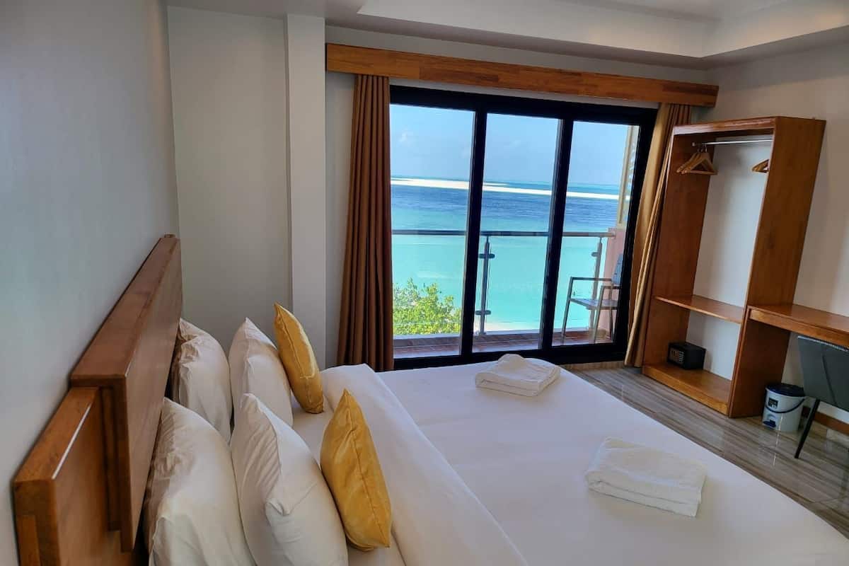 A hotel room with a large bed, a wooden headboard, a wardrobe, and a view of the ocean through a sliding glass door.