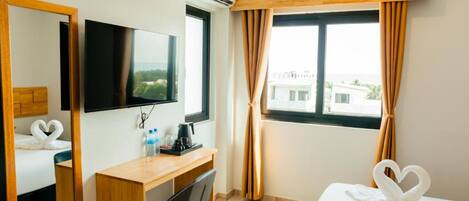 A hotel room with a bed, a desk with a chair, a flat-screen TV, a window with curtains, and a view of buildings outside.