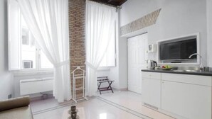 Una cocina luminosa con armarios blancos, un ventanal con cortinas traslúcidas, un televisor de pantalla plana y una mesita con un hervidor.