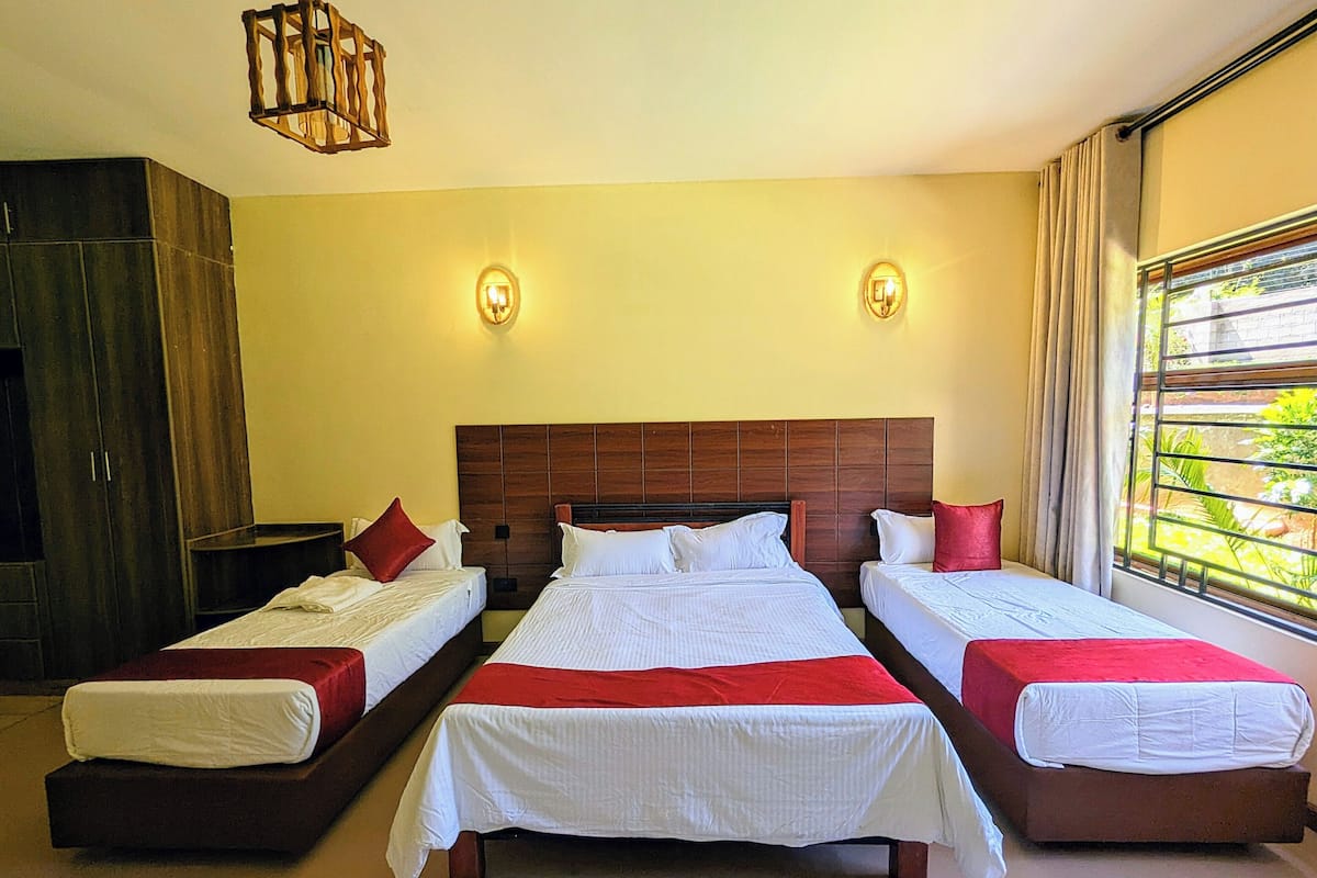 Habitación de hotel con tres camas dispuestas en fila, cabecera de madera, dos apliques de pared y un ventanal grande con cortinas.