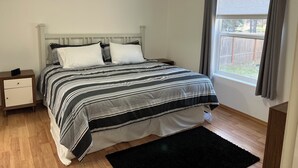 Une chambre à coucher comprenant un lit, une table de chevet, un ventilateur de plafond, une fenêtre avec des rideaux et une vue sur le jardin arrière.
