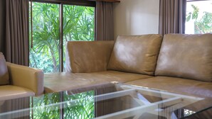 A living room with a beige leather sofa, a glass coffee table, and a view of green foliage through large windows. - Cozy gray garden (Pattaya)