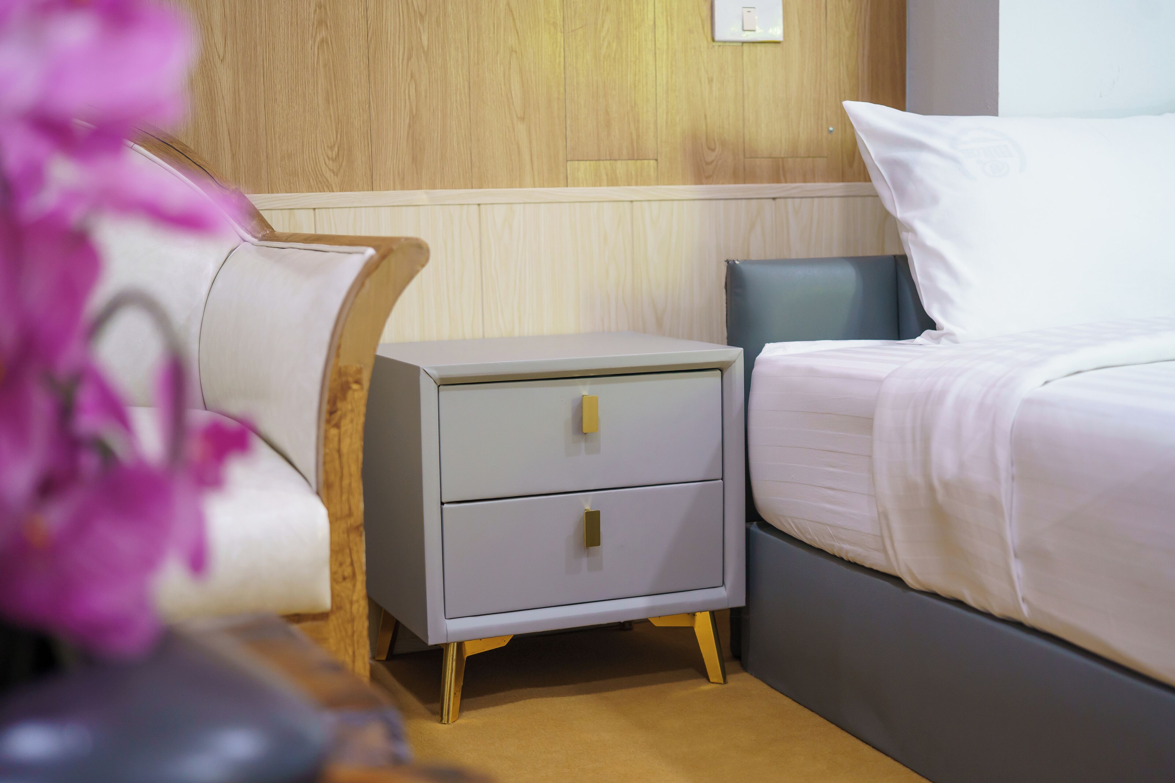 Un lit bien fait, avec du linge de lit blanc, une table de chevet grise dotée de deux tiroirs et une tête de lit en bois.
