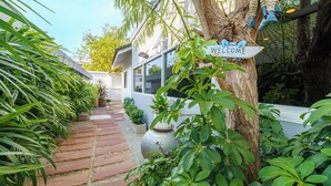 A pathway leading to a building with a 'Welcome' sign, surrounded by lush greenery and a large pot. - Cozy gray garden (Pattaya)