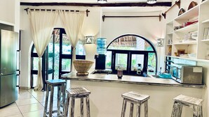 Una cocina con dispensador de agua, cafetera y un ventanal grande con cortinas. Se observan tres taburetes y un refrigerador.