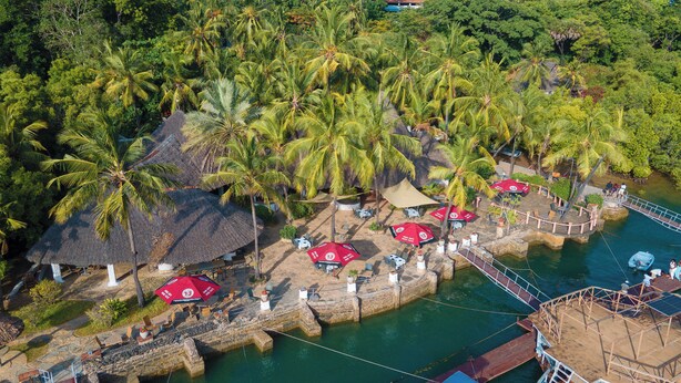 Vista aérea de un resort con cabañas de techo de paja, palmeras y un cuerpo de agua con un muelle de madera.