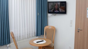 Une pièce avec une chaise et une table en bois, un téléviseur fixé au mur et une fenêtre ornée de rideaux bleus.