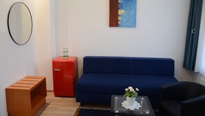 Une pièce avec un canapé bleu, une petite table sur laquelle est posé un vase de fleurs, un buffet rouge, un banc en bois et un miroir fixé au mur.
