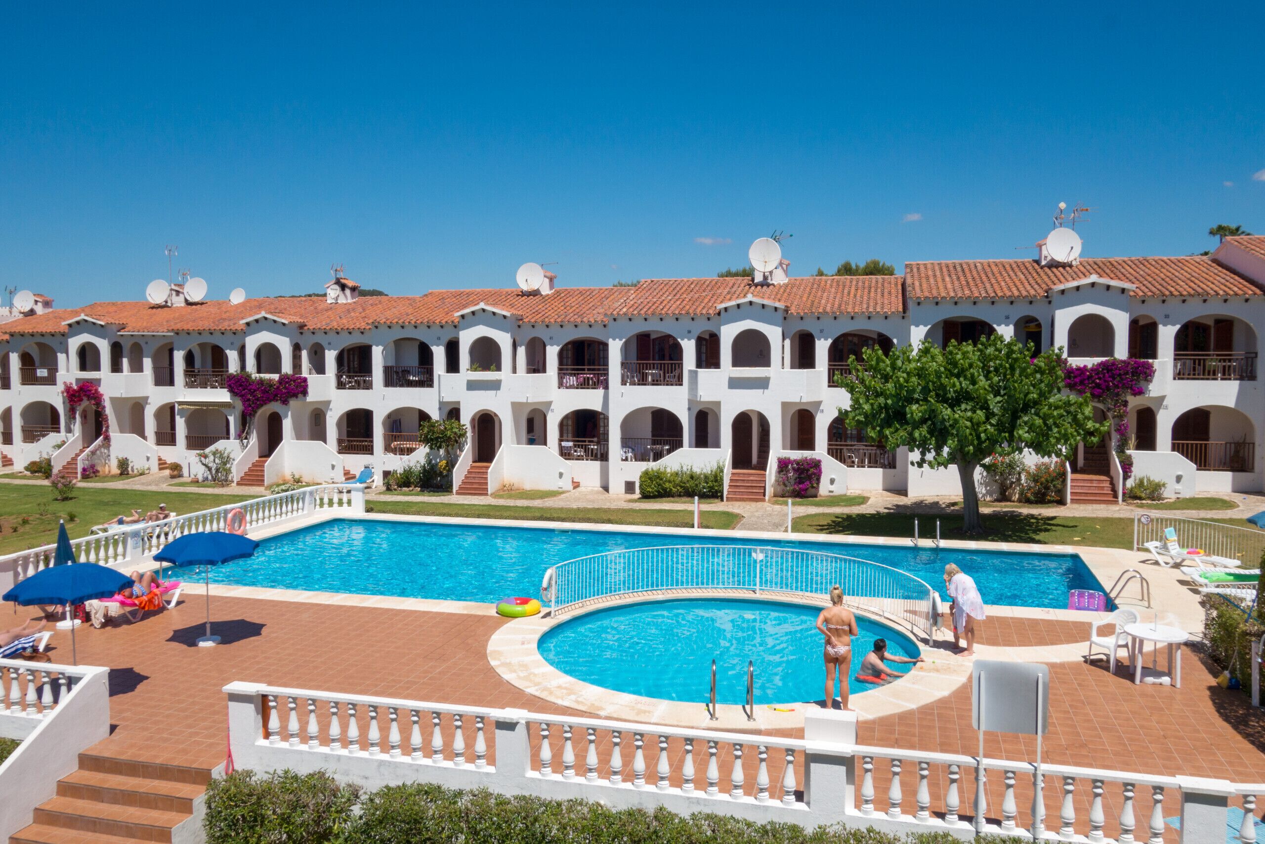A swimming pool surrounded by a white building with arched windows and a tiled roof.