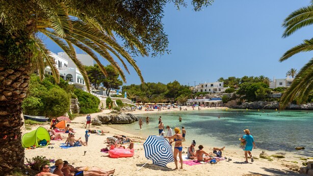 A beach with people sunbathing, a clear blue sea, and buildings in the background. - APARTAMENTO FONA (Cala Blanca)